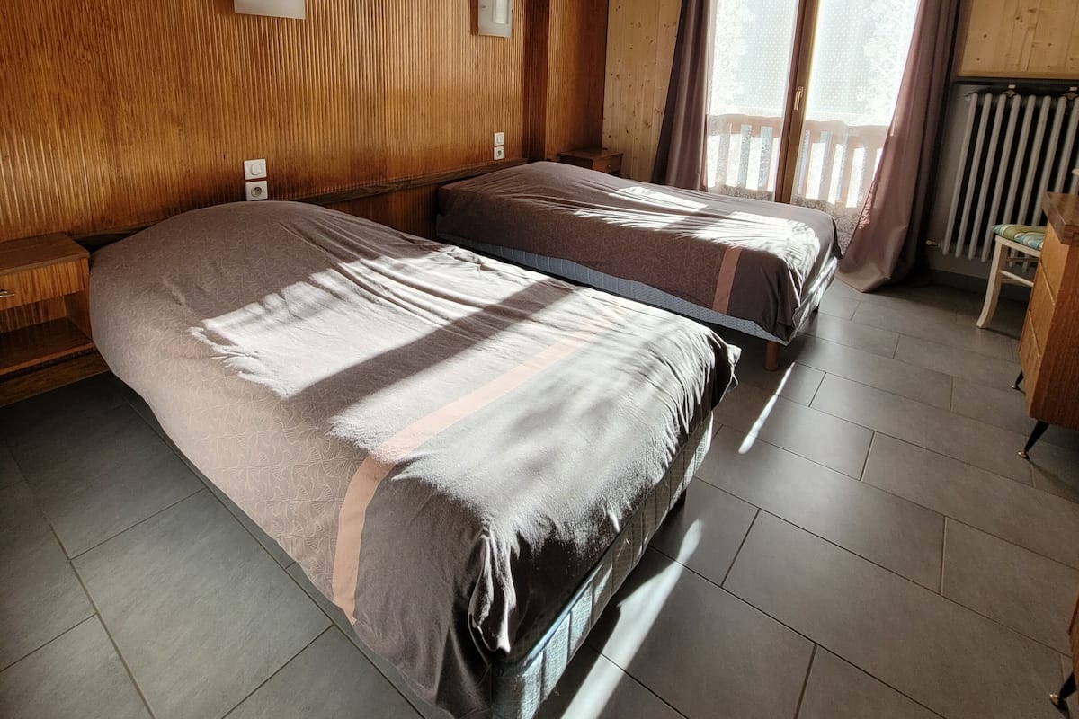 Una habitación con paneles de madera, dos camas, una mesita y una ventana con cortinas.
