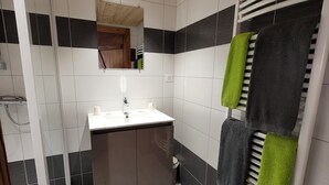 Een moderne badkamer met een wit wastafelmeubel, wandtegels in donkere en lichte tinten, een houten plafond en een handdoekrek met groene en zwarte handdoeken.