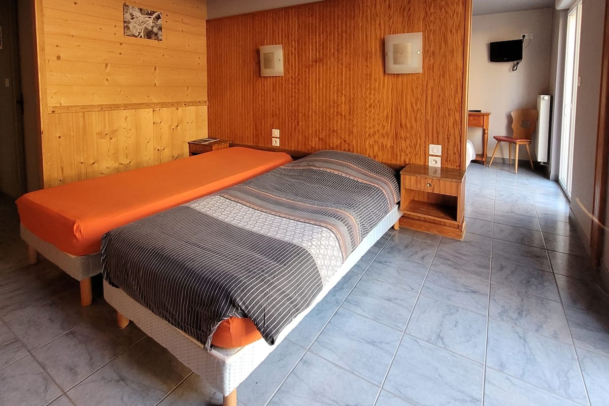 Un dormitorio con cama, paredes y puerta de madera, una mesita, una silla y un televisor fijado a la pared.