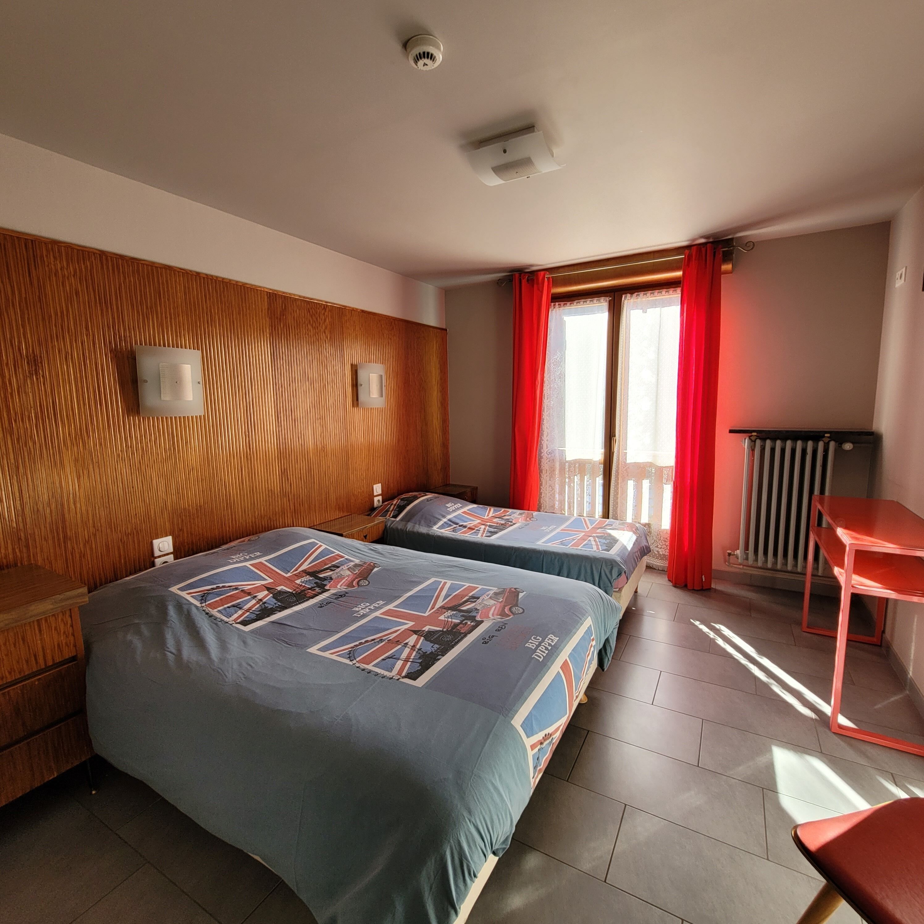 A hotel room with two beds, a wooden panel wall, a radiator, and a window with red curtains.