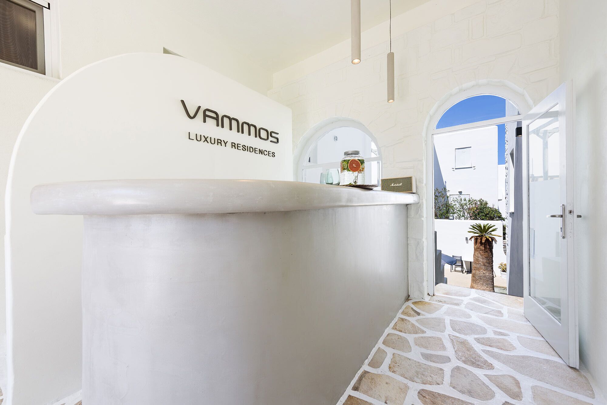 Ein weißer Empfangstresen mit der Beschriftung 'VAMMOS LUXURY RESIDENCES', ein Boden mit Steinplatten und Blick ins Freie.