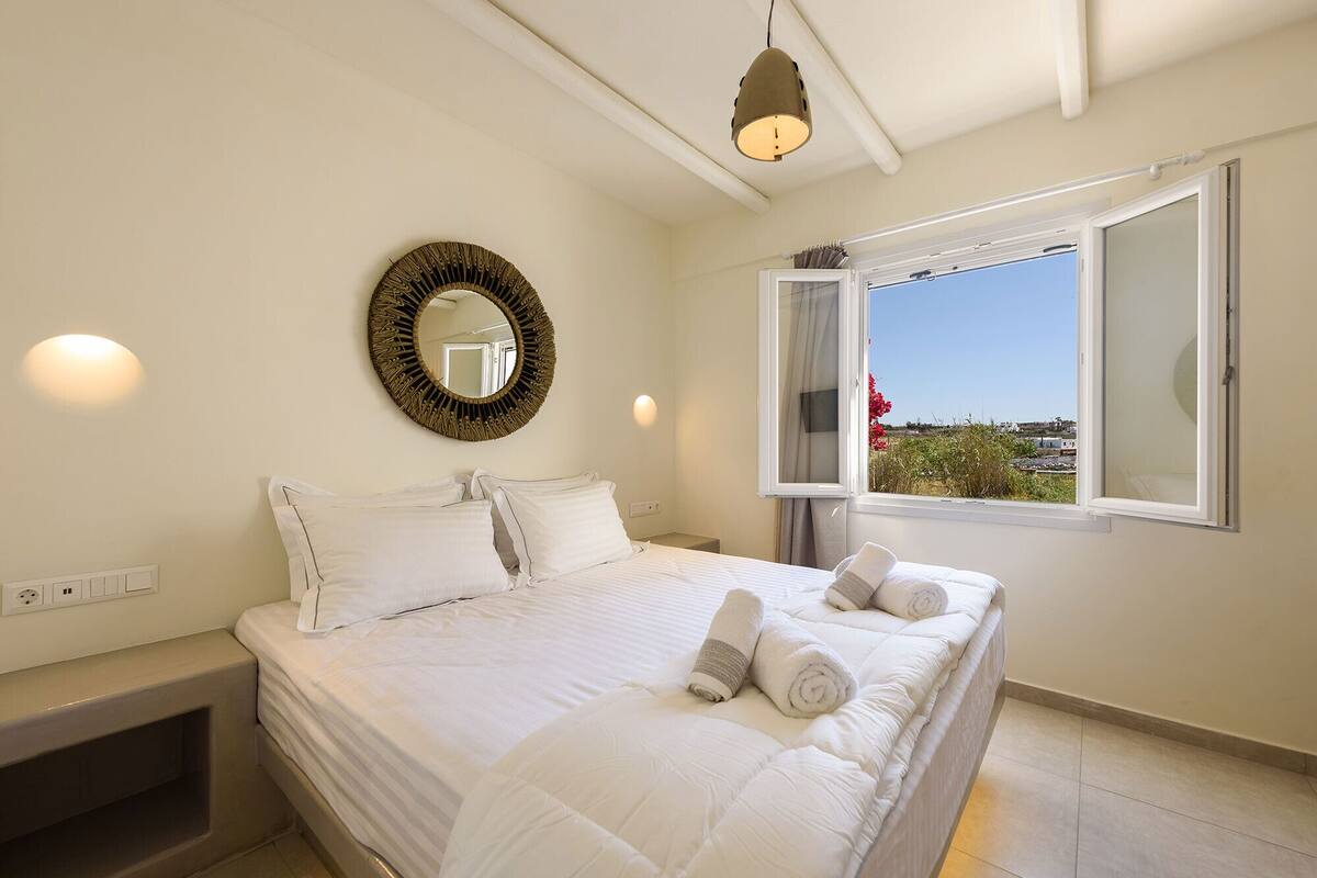 A bedroom with a bed, a round mirror, a window with curtains, and a view of the outside.