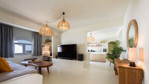 A modern living room with a sofa, coffee table, TV, and kitchen area with a dining table and chairs. - Vammos Luxury Apartments (Paros)