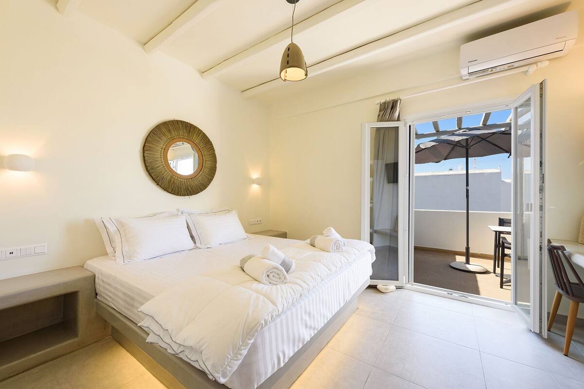 A bedroom with a bed, a round mirror, and a sliding glass door leading to an outdoor area with a table and chairs.