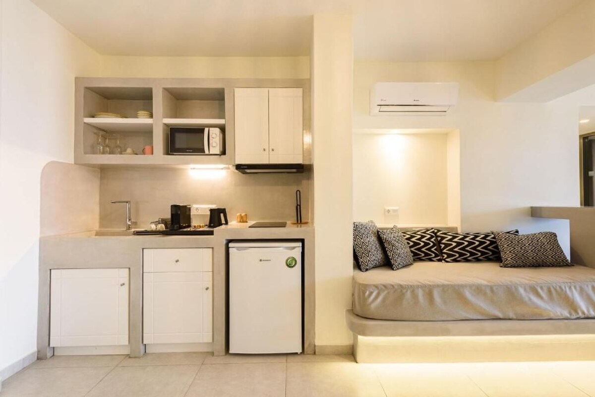 A compact kitchenette with a microwave, coffee maker, and a small refrigerator. There is a sofa with patterned cushions and an air conditioning unit mounted on the wall.