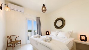 A bedroom with a bed, a chair, a bedside table, a mirror, and a view of the outdoors. - Vammos Luxury Apartments (Paros)