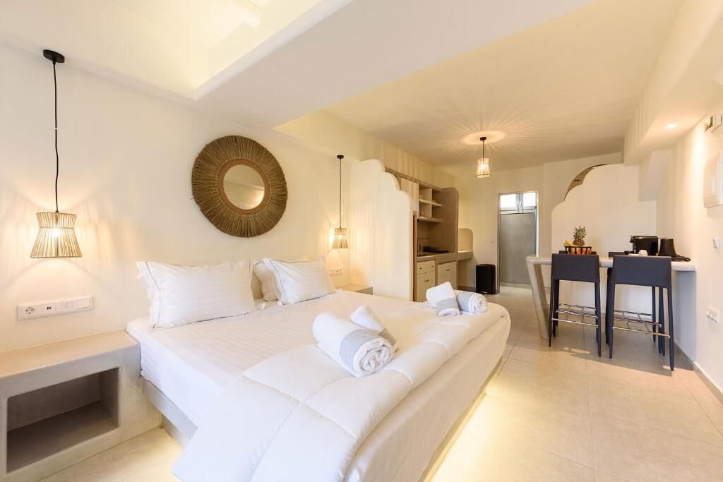 A hotel room with a large bed, bedside tables, a round mirror, and a kitchenette area.