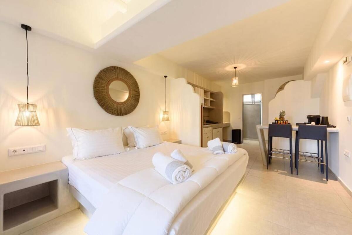 A hotel room with a large bed, bedside tables, a round mirror, and a kitchenette area.