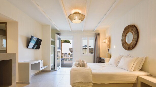 A modern bedroom with a bed, a TV, and a view of the outdoor area. - Vammos Luxury Apartments (Paros)