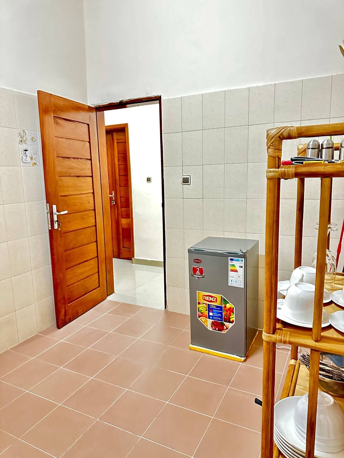 A room with a wooden door, a small refrigerator, and a bamboo shelf with dishes.
