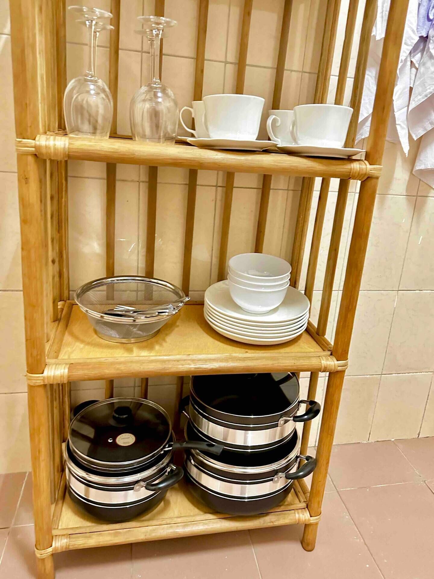 En hylla av bambu med köksredskap, inklusive en glasskål, vita koppar och staplade tallrikar.