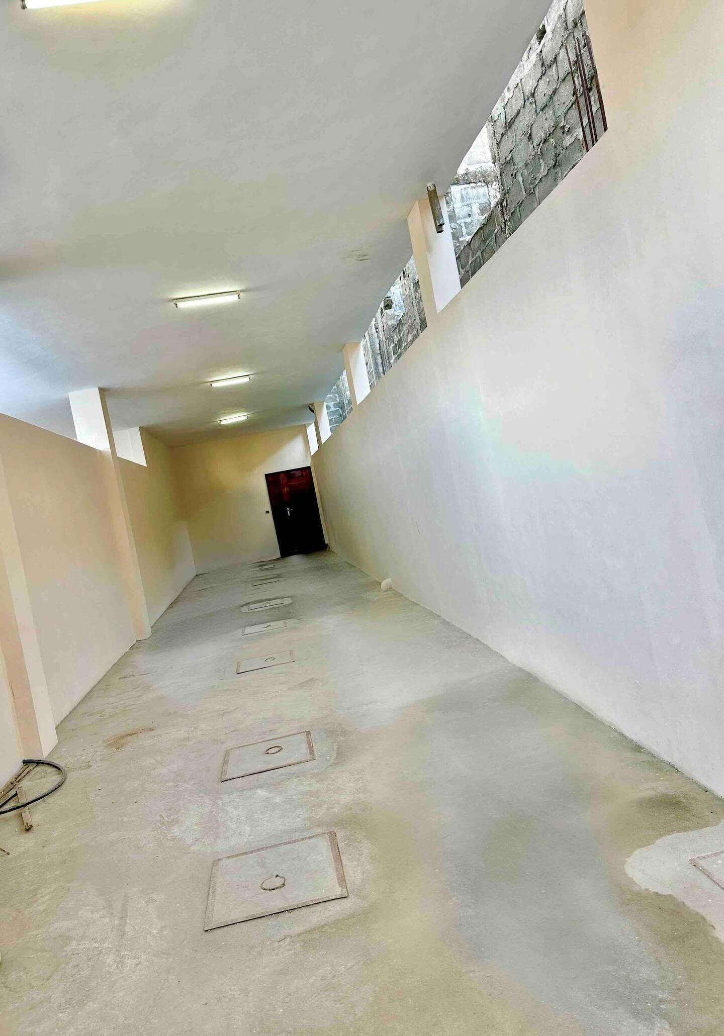 A narrow, unfinished hallway with exposed concrete floors and walls, a dark door at the end, and a small window with a brick pattern on the right wall.