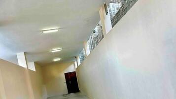 A narrow, unfinished hallway with exposed concrete floors and walls, a dark door at the end, and a small window with a brick pattern on the right wall.