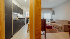 A modern kitchen with dark cabinets and a wooden stool, a beige sofa, and a red bean bag in a living area with a large window. - Good Morning Apartment With Garage by Vacationy (Porto)