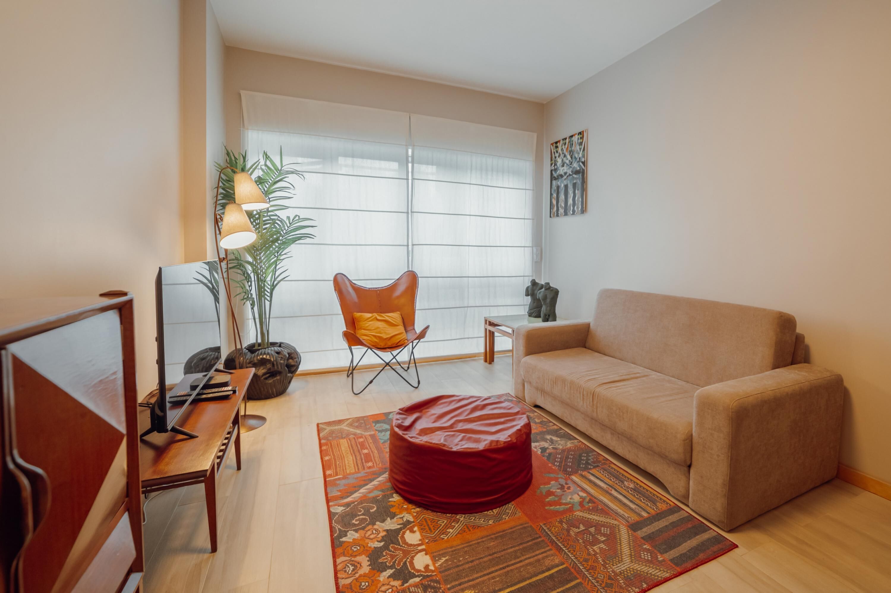 A modern living room with a sofa, a red bean bag, a wooden coffee table, a floor lamp, and a large window with sheer curtains.