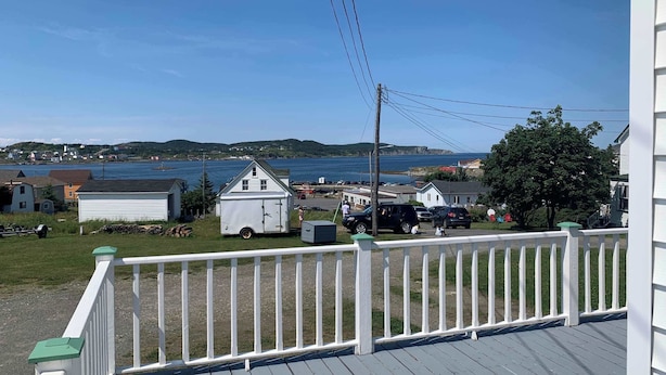 A view from a balcony with a white railing, overlooking a small town with houses, a trailer, and a body of water.