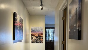 A hallway with wooden flooring, white walls, and ceiling lights. There are three framed artworks on the walls and a door on the right. - Cozy 2-Bedroom Upstairs Apartment in  Paradise (Paradise)
