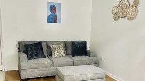 A living room with a grey sectional sofa, a matching ottoman, a white table, and a wall with decorative plates.
