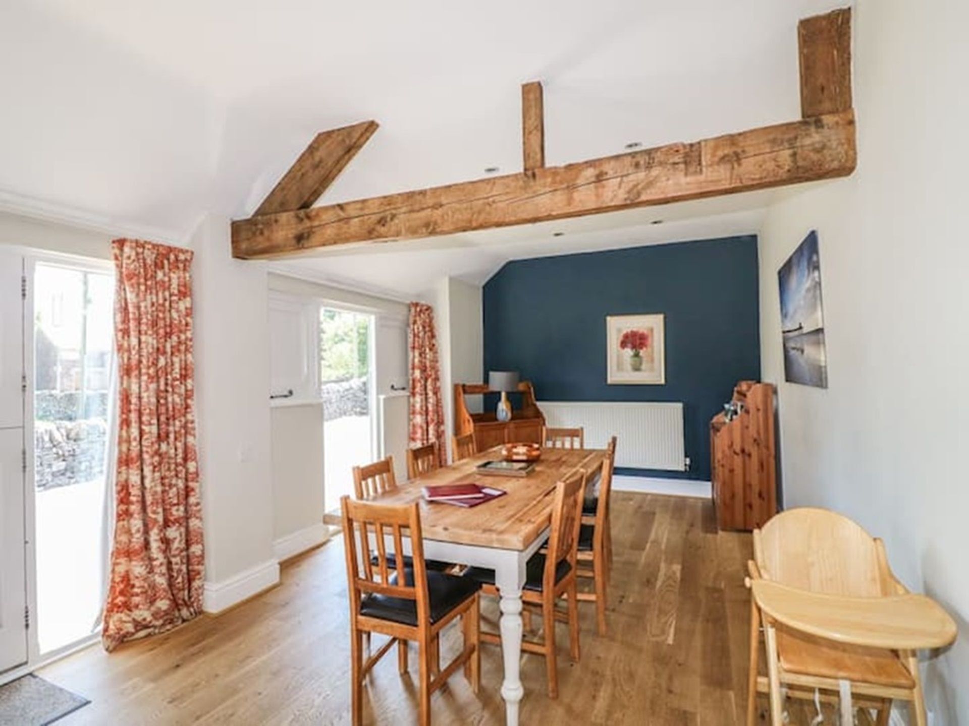 A dining area with a wooden table and chairs, a high ceiling with exposed beams, and a view of the outdoors through a door.