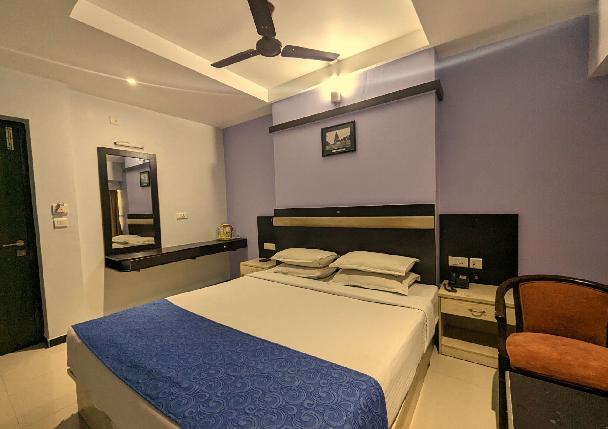 A hotel room with a bed, a nightstand, a chair, a ceiling fan, and a wall-mounted picture.