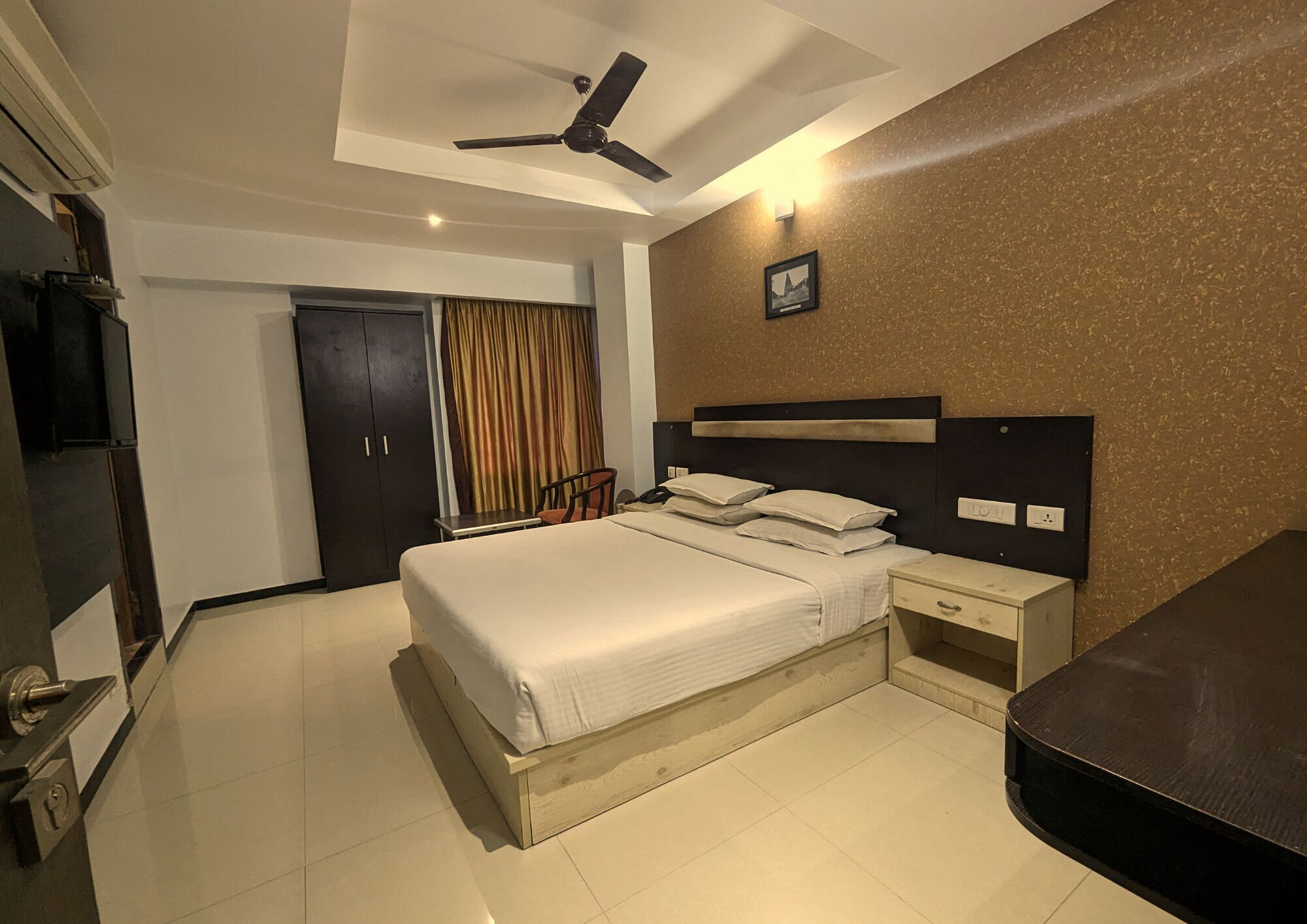 A hotel room with a bed, bedside tables, a desk, a chair, a ceiling fan, a door, and a wall-mounted light.