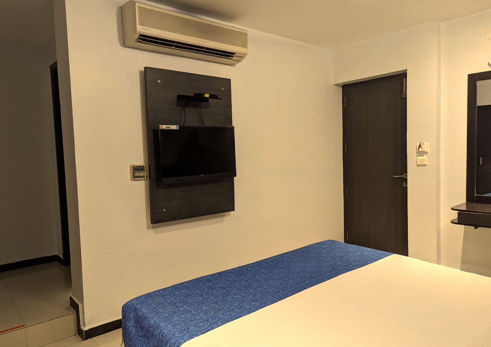 A hotel room with a bed, a wall-mounted air conditioner, a door, and a small shelf.