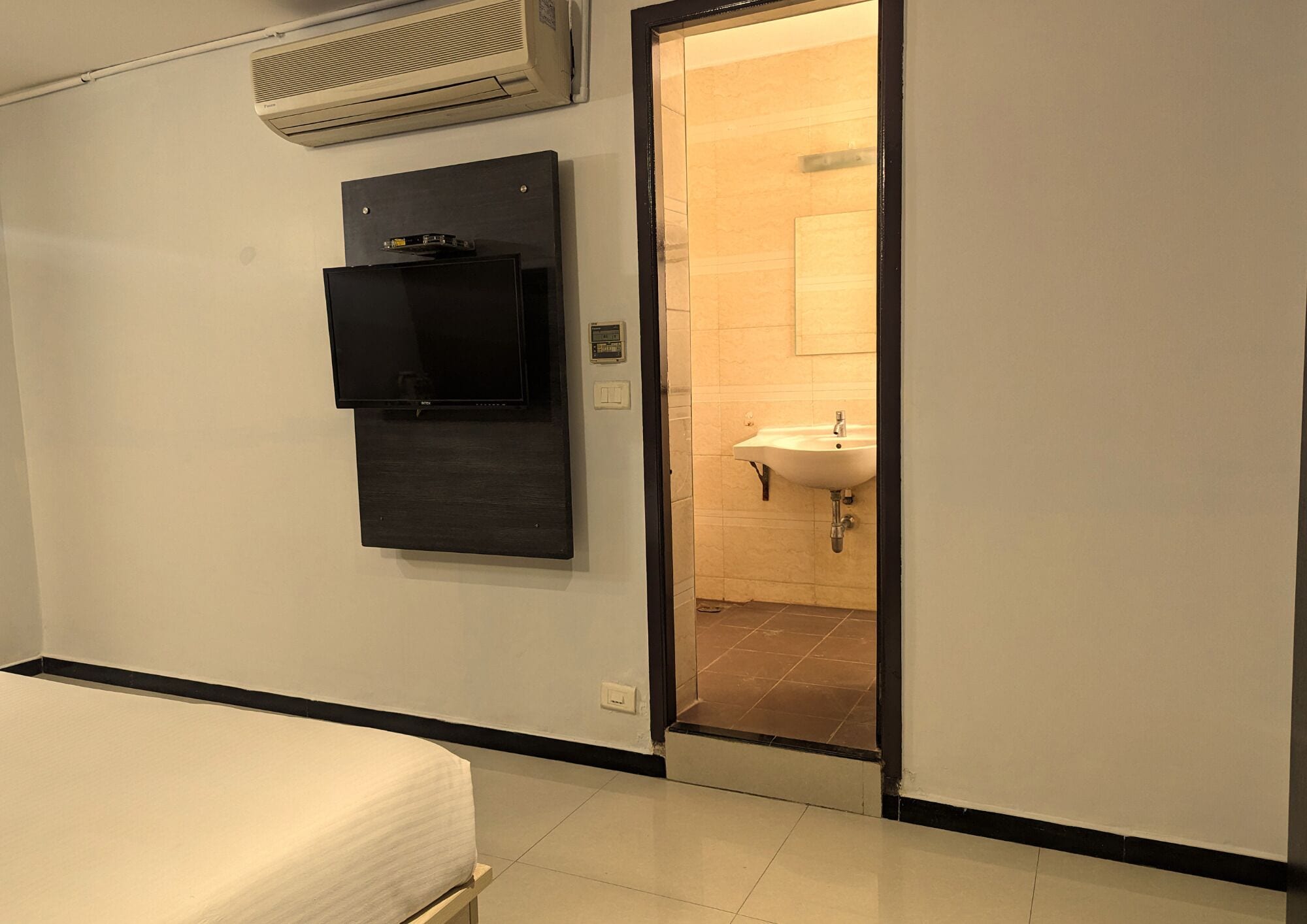 Habitación de hotel con cama, televisión montada en la pared, aire acondicionado instalado y baño visible a través de una puerta abierta.