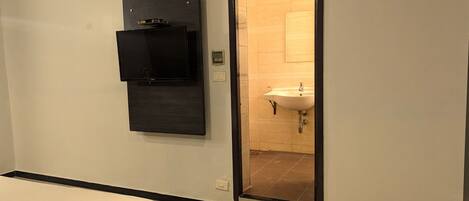 Habitación de hotel con cama, televisión montada en la pared, aire acondicionado instalado y baño visible a través de una puerta abierta.