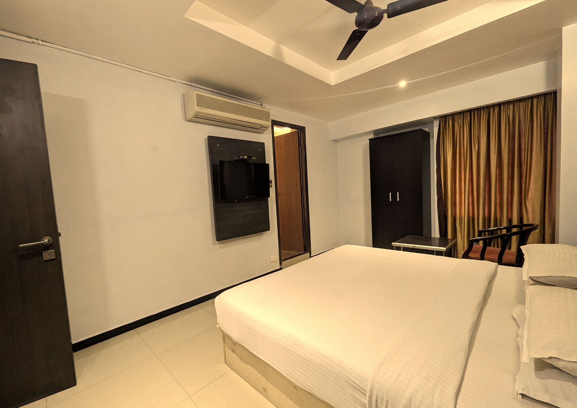 A hotel room with a bed, a desk, a chair, a wardrobe, a TV, and an air conditioning unit.