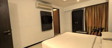 A hotel room with a bed, a desk, a chair, a wardrobe, a TV, and an air conditioning unit.
