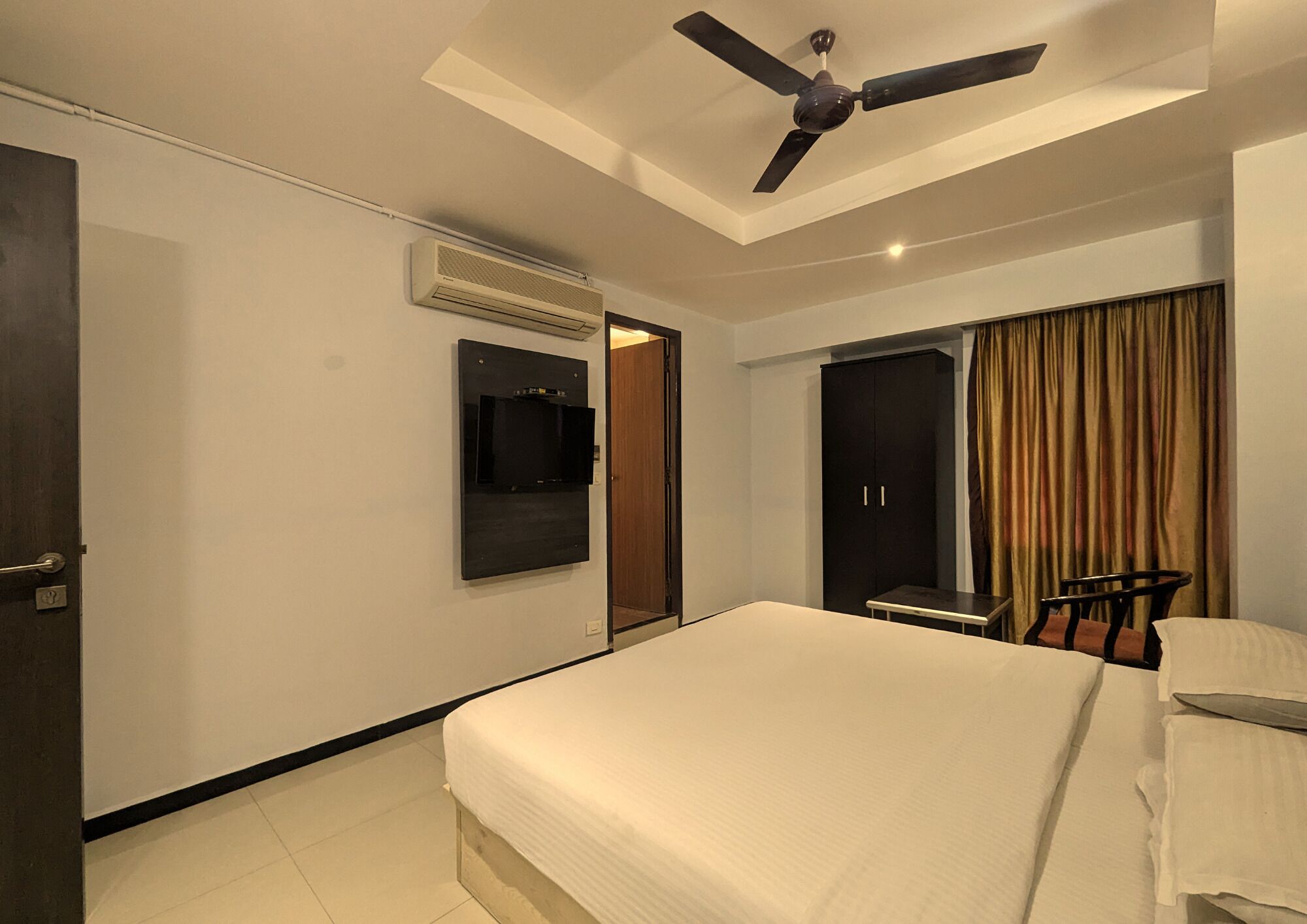 Ein Hotelzimmer mit einem Bett, einem Deckenventilator, einer Klimaanlage, einem an der Wand montierten Fernseher, einem Kleiderschrank und einem Fenster mit Vorhängen.
