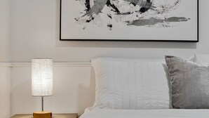 A neatly made bed with white and gray bedding, a bedside lamp on a wooden nightstand, and a framed abstract painting on the wall. - Enjoy a stylish experience at this fresh and clean  lower apartment!  This place is newly renovated and perfect for a stay in the Metro Detroit. It's located near the Detroit Zoo right by major highways. This home is a duplex building and includes 2 (Oak Park)