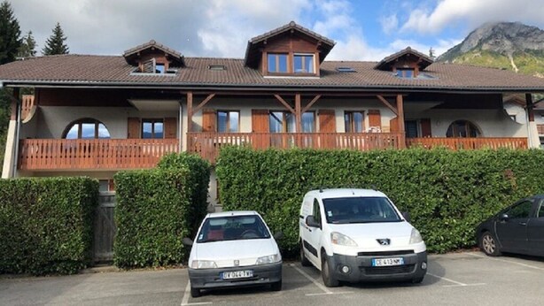 Un bâtiment doté d’un balcon en bois, un parking où sont garées deux voitures, et une montagne en arrière-plan.