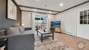 A living room with a grey sofa, a coffee table, a patterned rug, a painting on the wall, and a sliding glass door leading to an outdoor area.