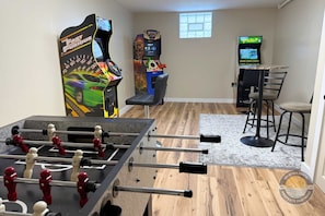 Una habitación con mesa de fútbol, dos juegos de arcade, una zona de bar con taburetes y un ventanal que proporciona luz natural.