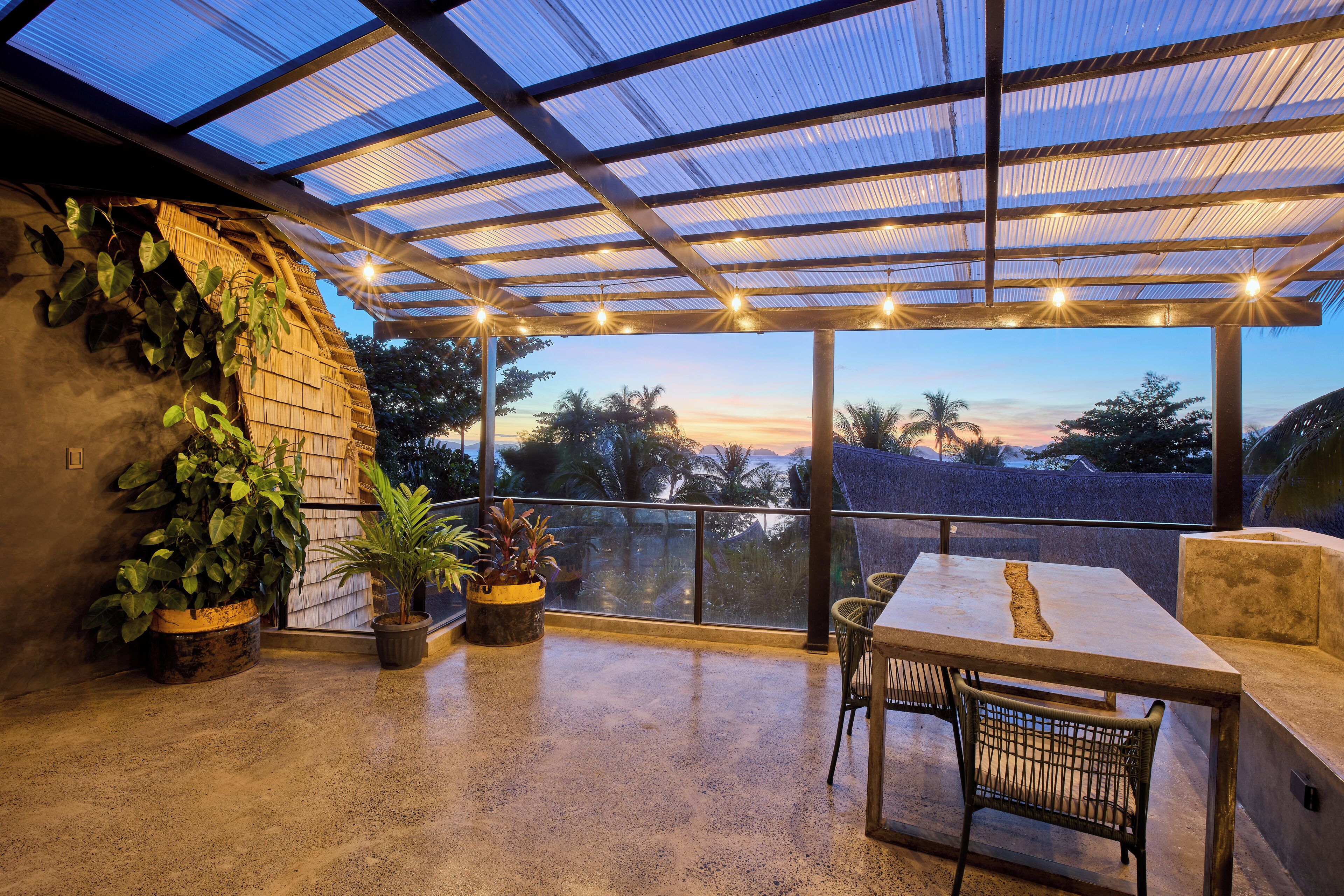 Un espacio al aire libre techado con una mesa y sillas, plantas en macetas, y vistas a palmeras y un atardecer.