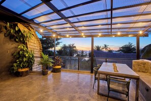 A covered outdoor area with a table and chairs, potted plants, and a view of palm trees and a sunset.