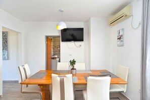 Un coin repas avec une table en bois, des chaises blanches, une plante en pot et un téléviseur fixé au mur.