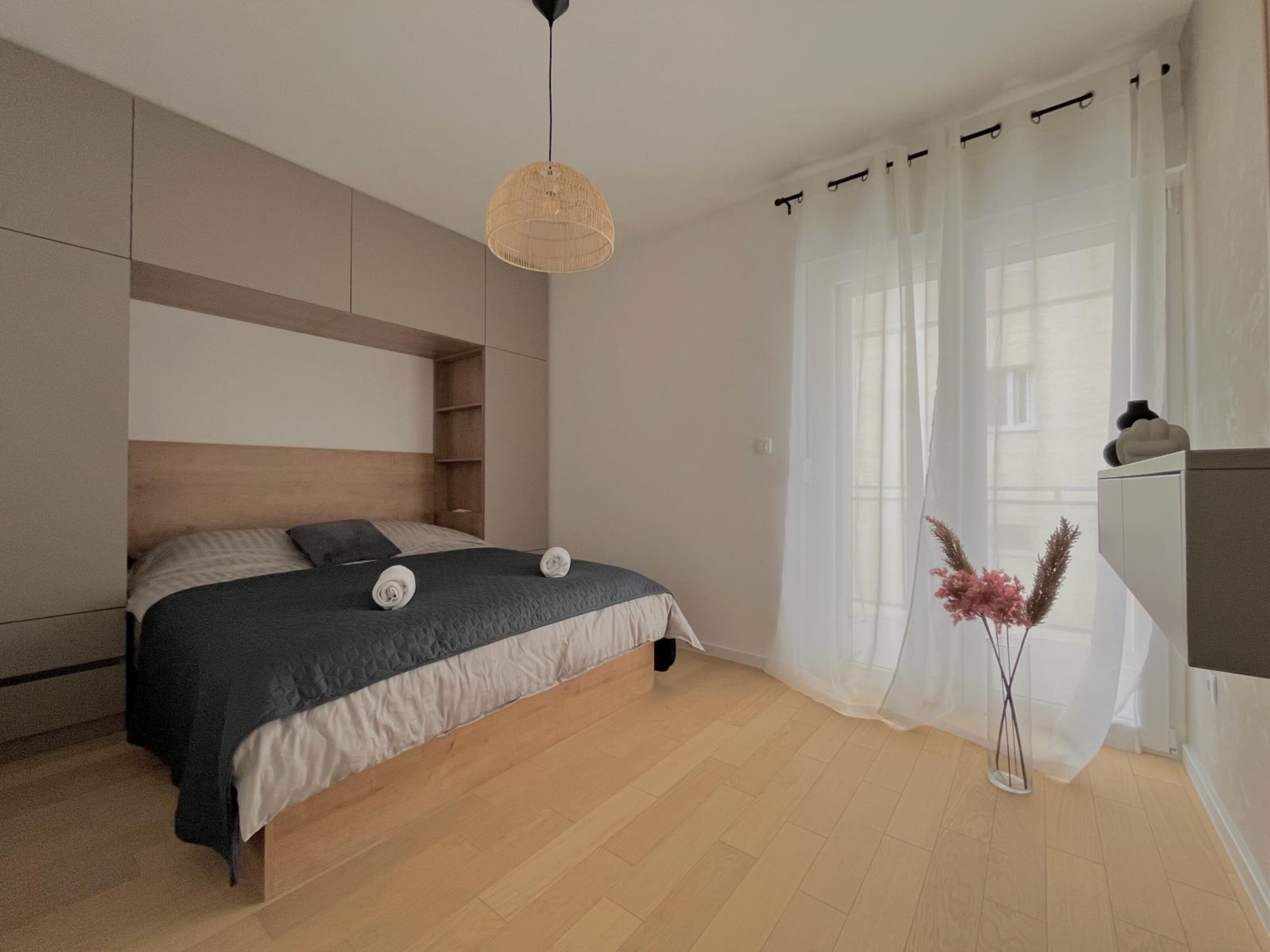 A modern bedroom with a bed, a hanging light fixture, a window with sheer curtains, and a small bedside table with a vase.