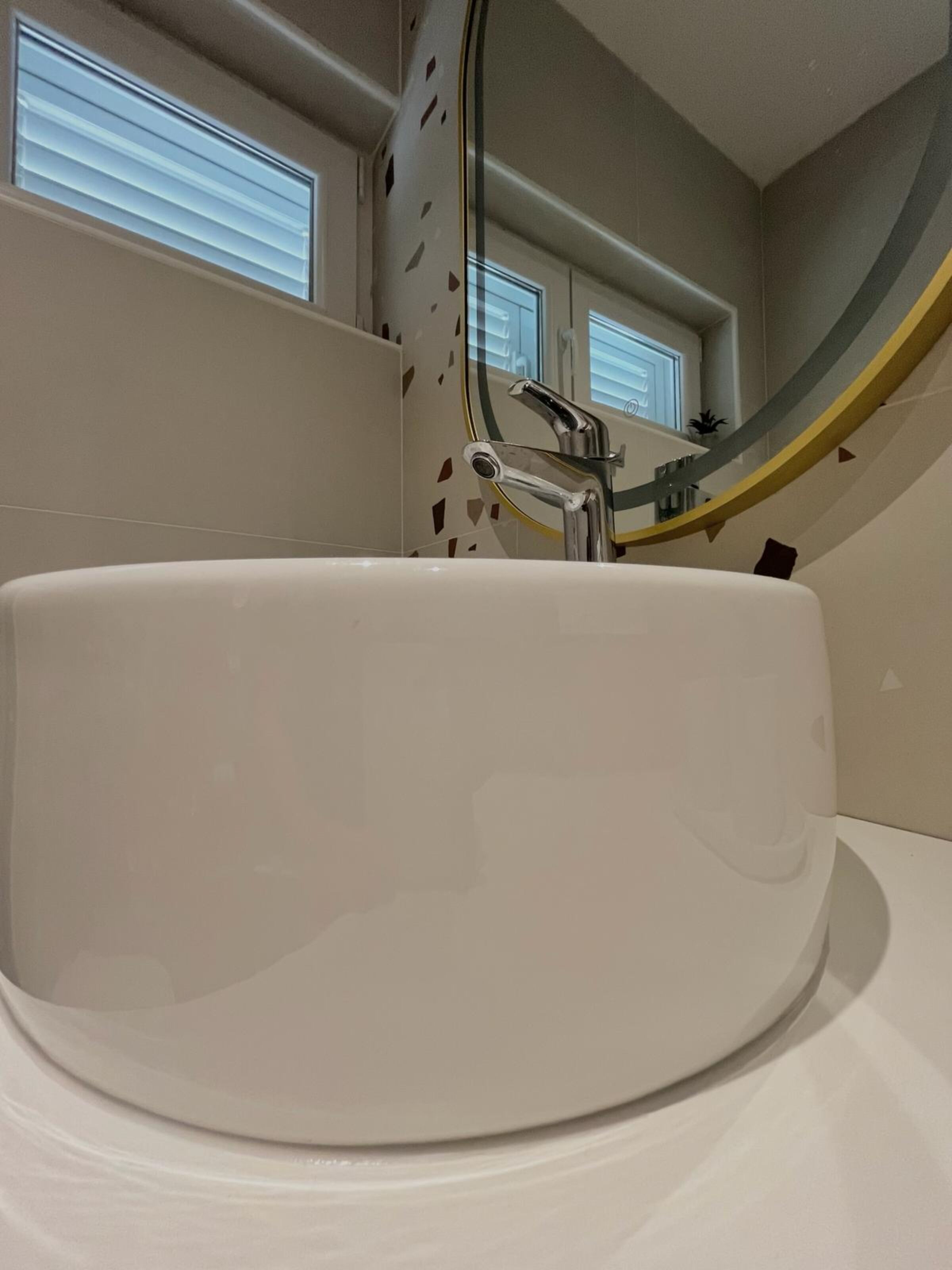 A modern bathroom with a large white sink, a curved mirror, and a window with blinds.