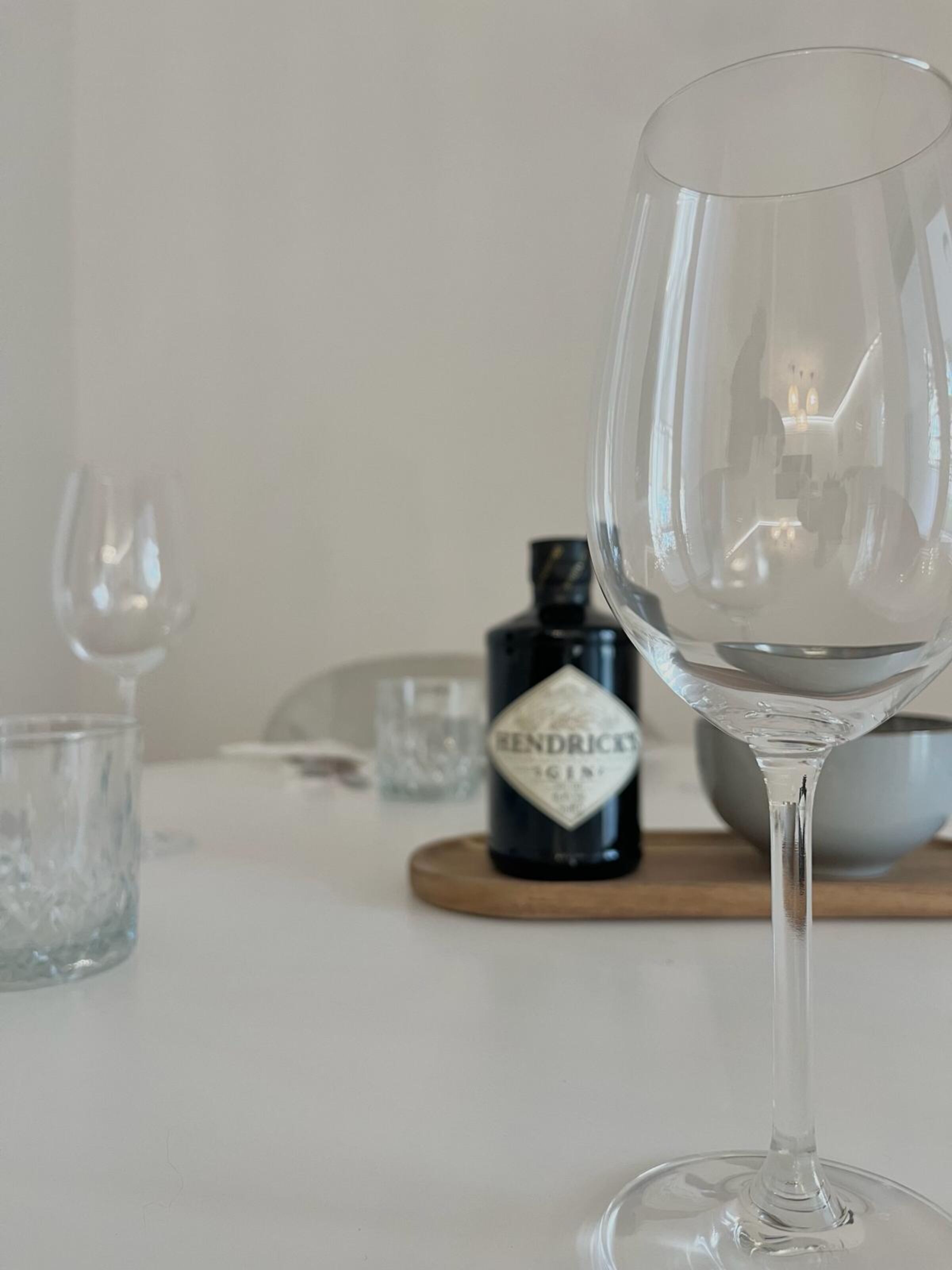 A clear glass wine glass on a table with a bottle labeled "HENDRICK'S" and a bowl in the background.