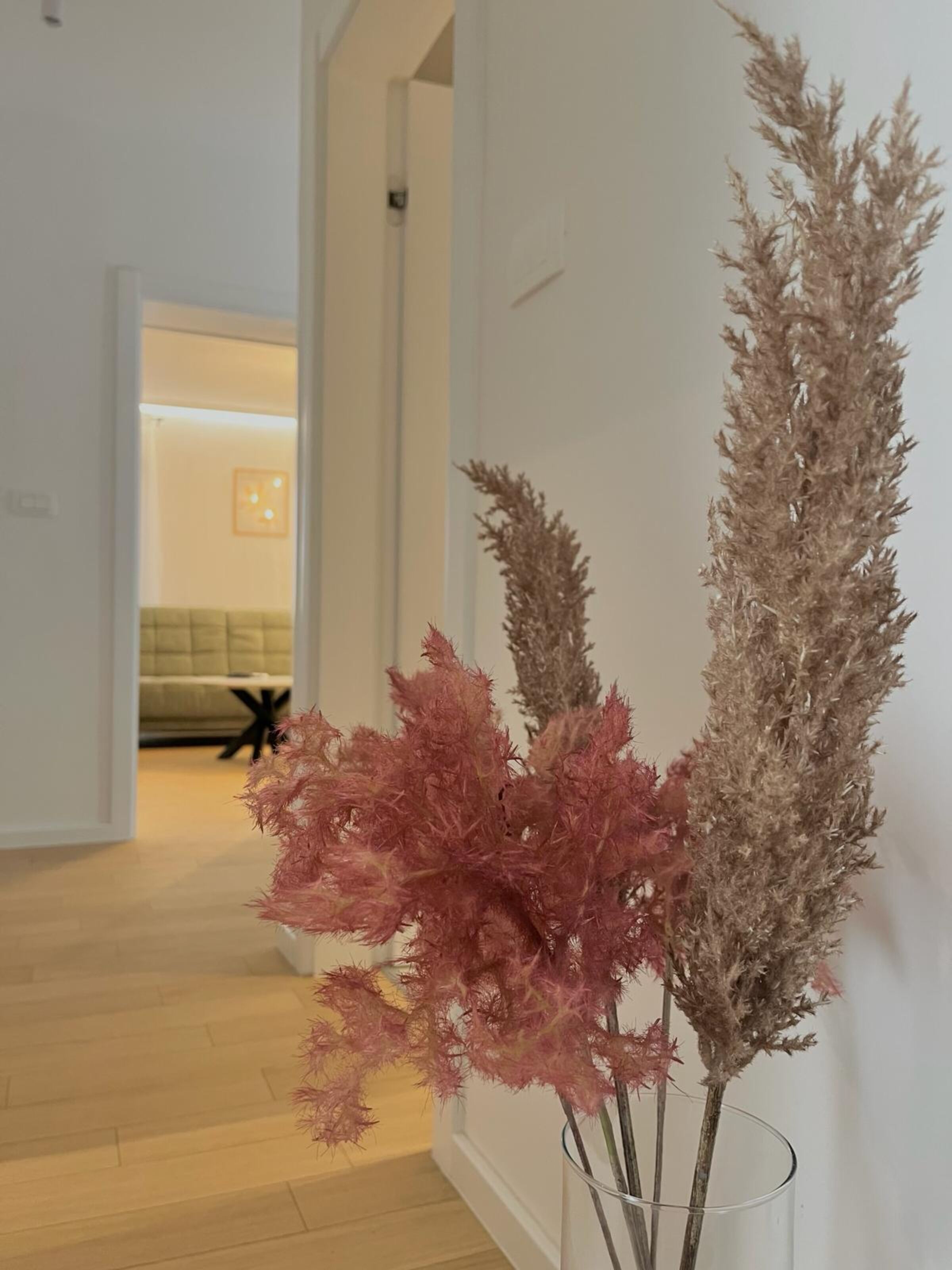Un couloir d’où l’on aperçoit une pièce au fond, un plancher en bois et un vase contenant des plantes séchées, le tout sur un mur blanc.
