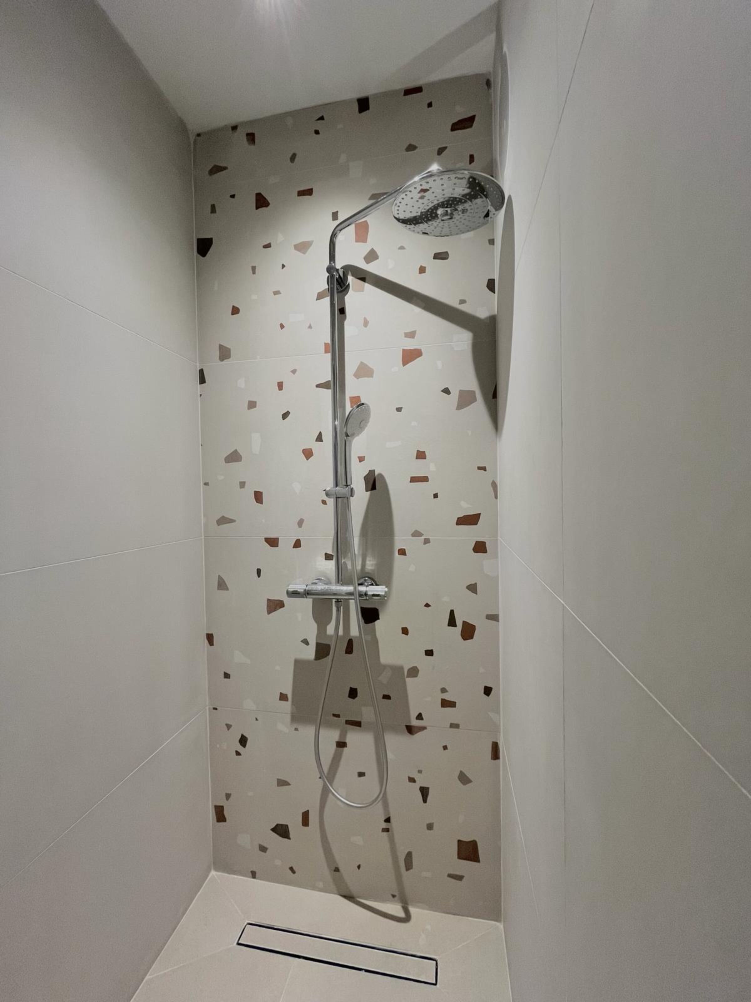 A shower with a rain showerhead and handheld showerhead, set against a wall with a terrazzo tile pattern.