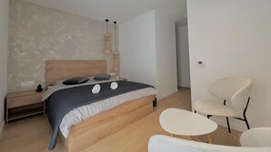 Un dormitorio moderno con una cama de madera, una mesita de noche pequeña, una lámpara de noche, una silla y una pequeña mesa.