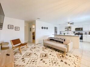 A modern living room with a sofa, a wooden chair, and a patterned rug. A kitchen with white cabinets and appliances is visible in the background.