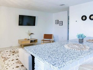 A modern living room with a flat-screen TV mounted on the wall, a wooden cabinet, a patterned rug, a chair, and a decorative plant. - Fillmore Unit 14 | 1BR/1BA (Cape Canaveral)