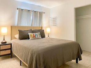 A neatly made bed with a beige comforter and pillows, two bedside lamps, a window with curtains, and a door. - Fillmore Unit 14 (Cape Canaveral)