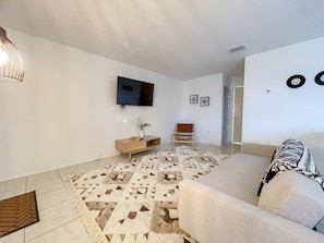 A modern living room with a flat-screen TV mounted on the wall, a patterned rug, a sofa, a wooden side table with a vase, and a bench. - Fillmore Unit 14 (Cape Canaveral)