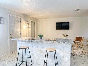 A kitchen with a white brick backsplash, two bar stools, a small island with a countertop, and a television mounted on the wall. - Fillmore Unit 14 (Cape Canaveral)