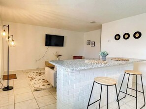 A kitchen with a white brick island, two bar stools, a flat-screen TV, and a floor lamp. - Fillmore Unit 14 | 1BR/1BA (Cape Canaveral)